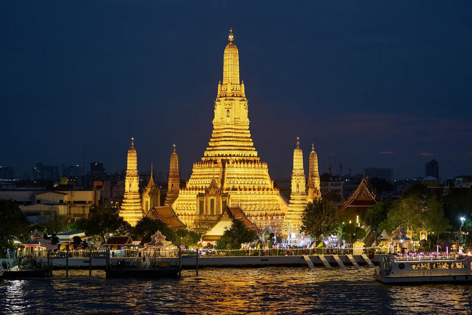 Wat Arun lit gold at night across the Chao Phraya River, with a lit-up boat passing