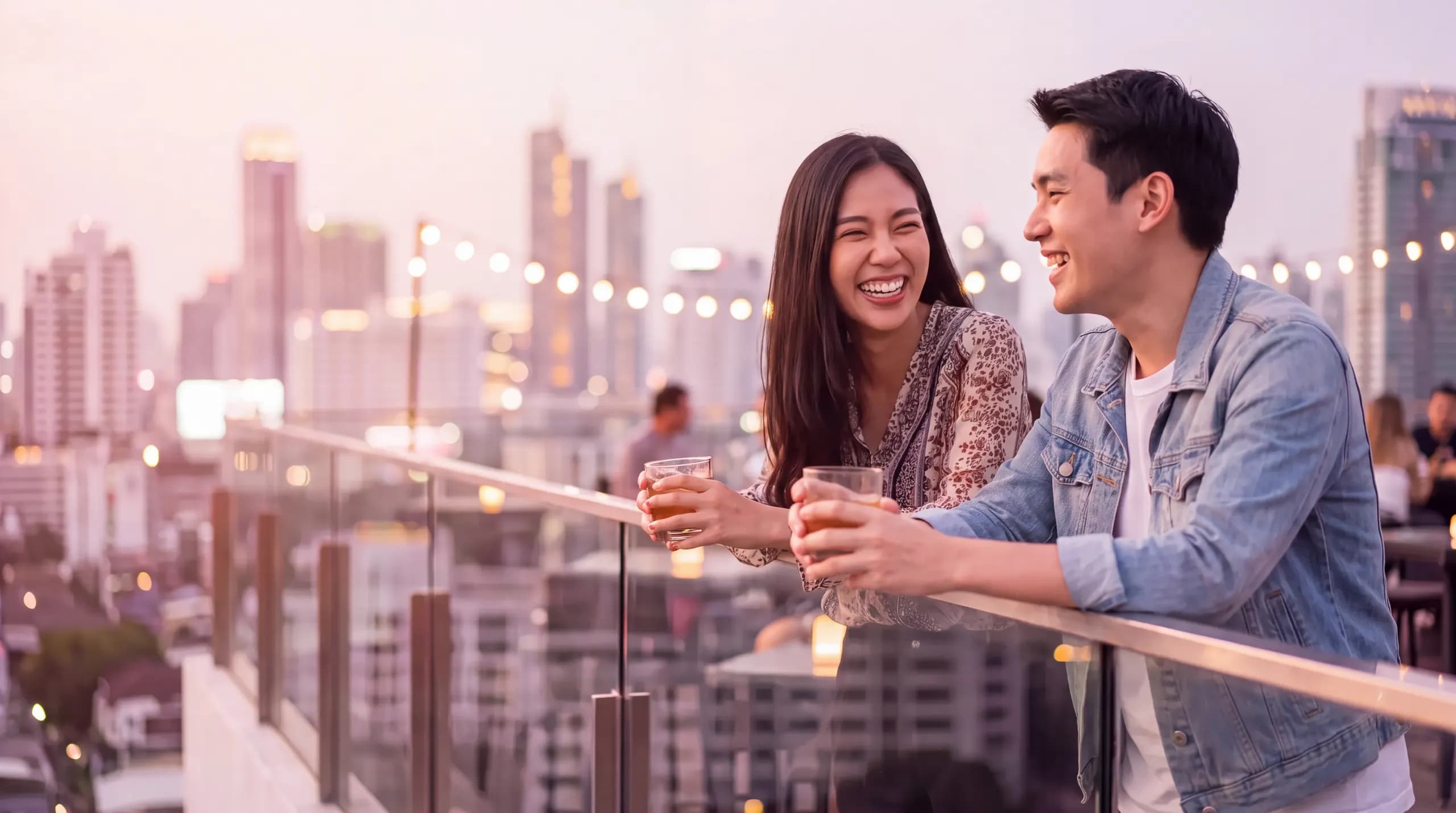 A Thai friend and a visitor laughing over drinks at a Bangkok rooftop bar at dusk
