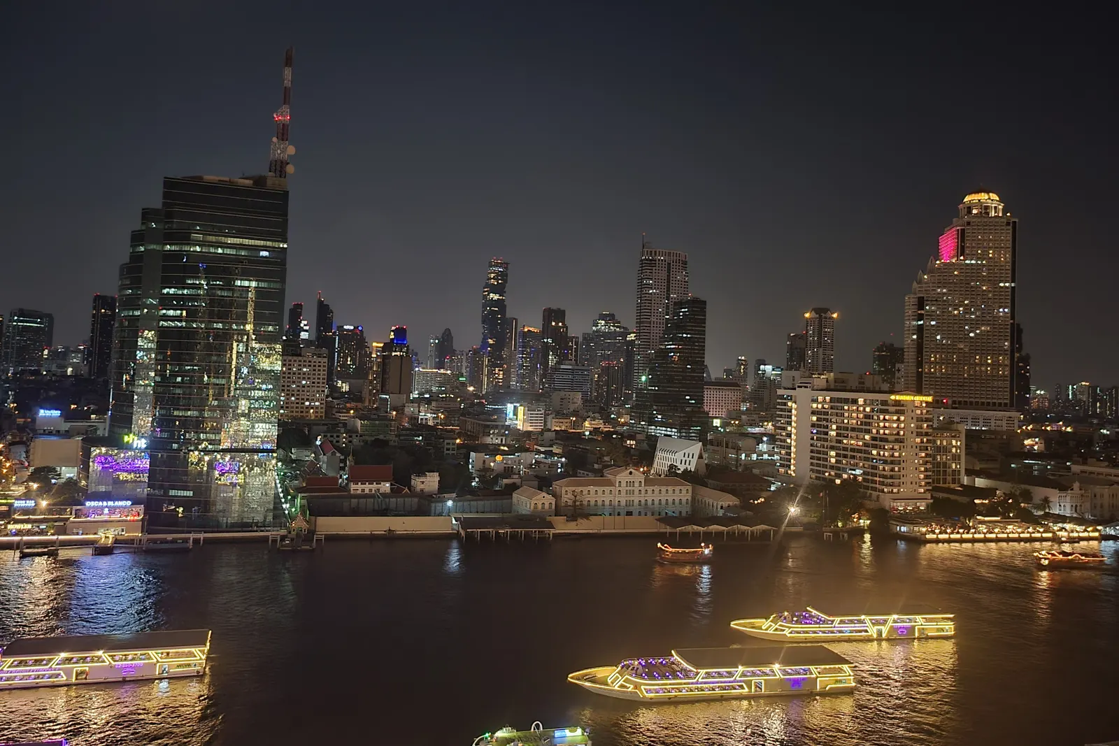 Lit-up dinner boats on the Chao Phraya River at night, with the Bang Rak towers behind
