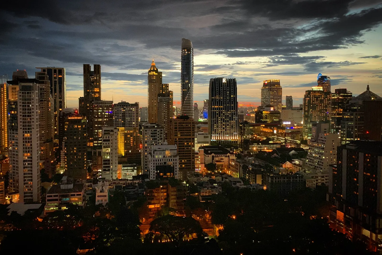Bangkok's towers lighting up at dusk under a cloudy sky, seen from a high floor