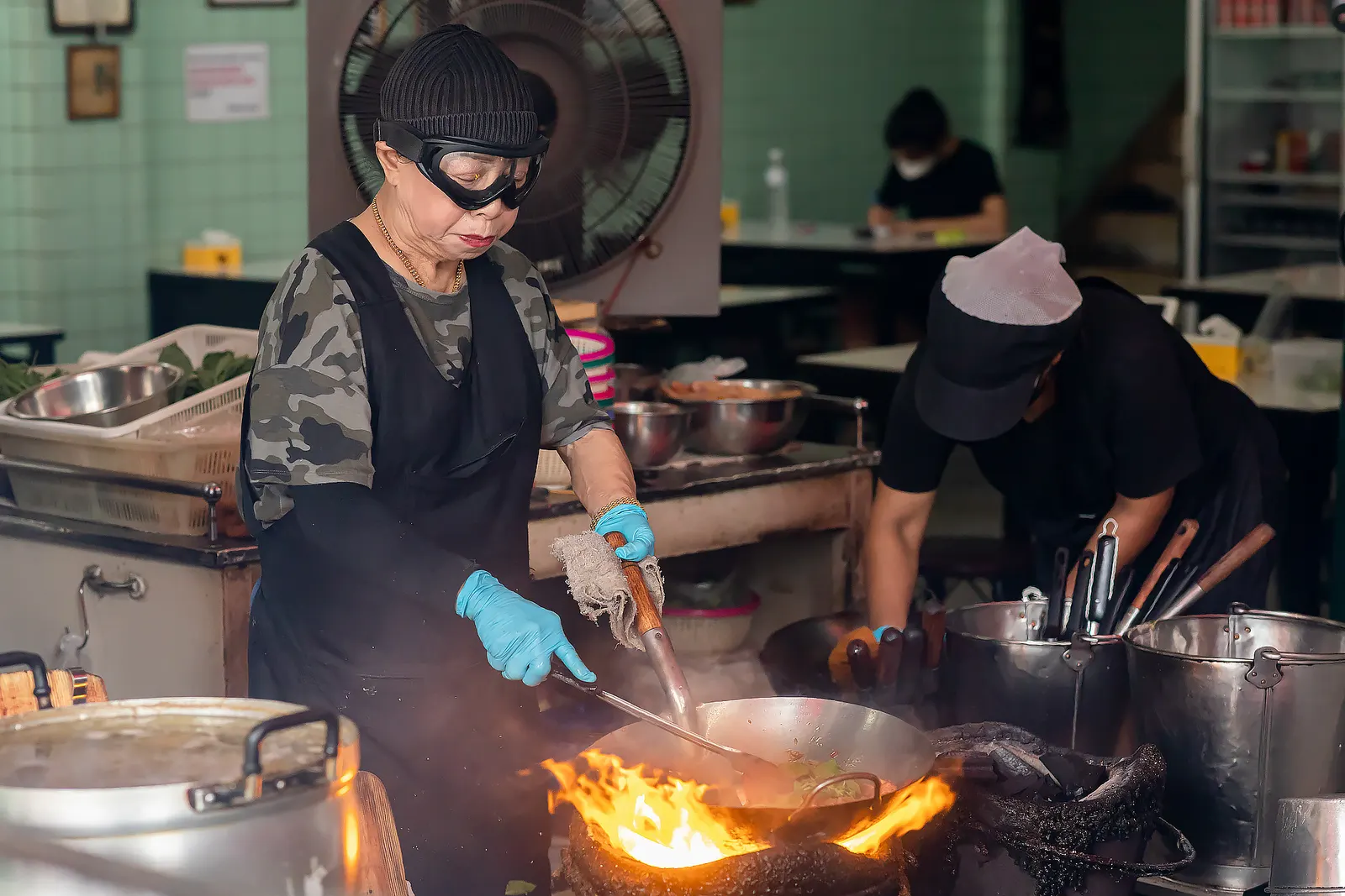Jay Fai in her ski goggles stirring a wok over a charcoal fire at her shop on Maha Chai Road