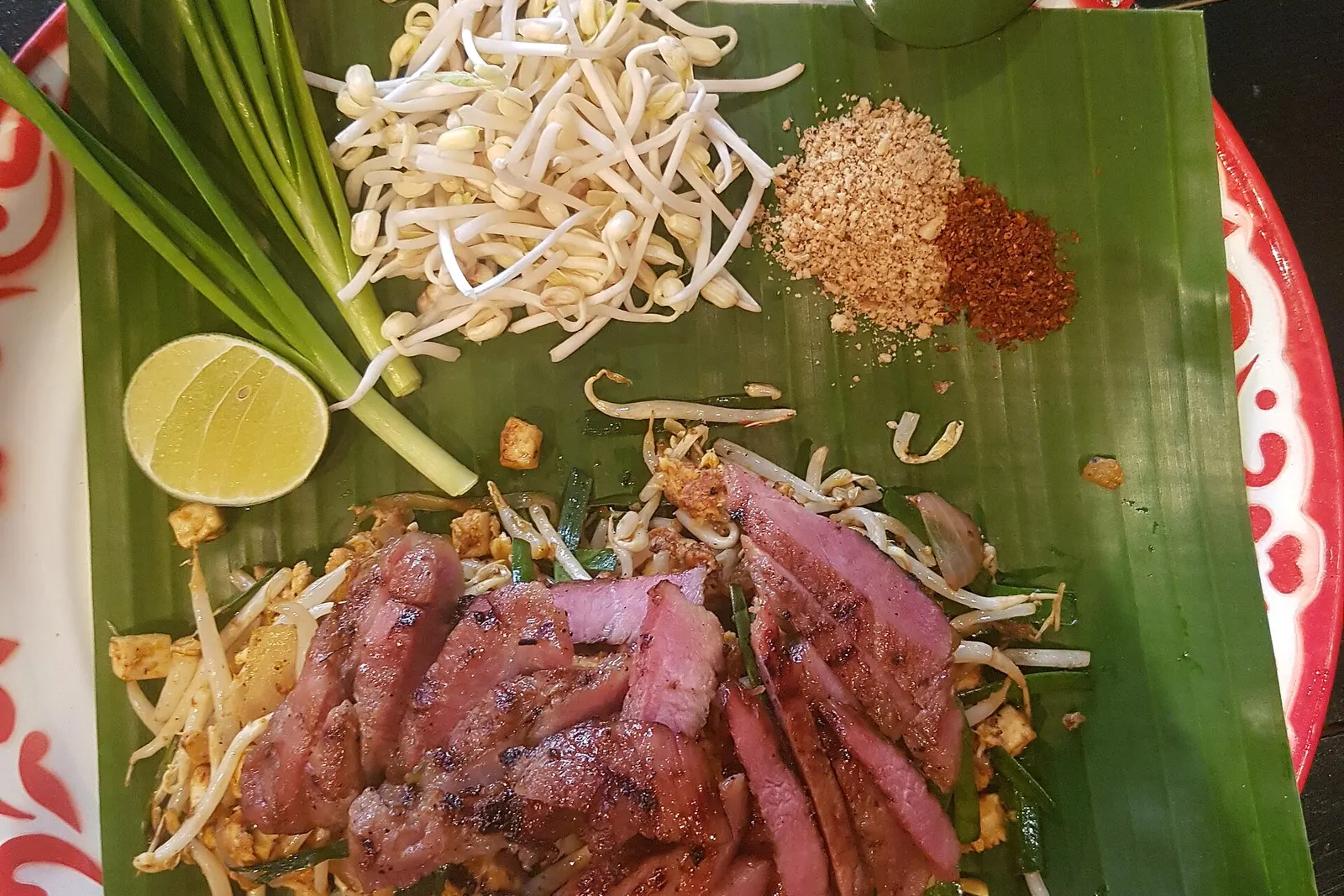 Pad thai with grilled pork on a banana leaf, with Chinese chives, half a lime, bean sprouts, ground peanuts and chilli flakes on the side