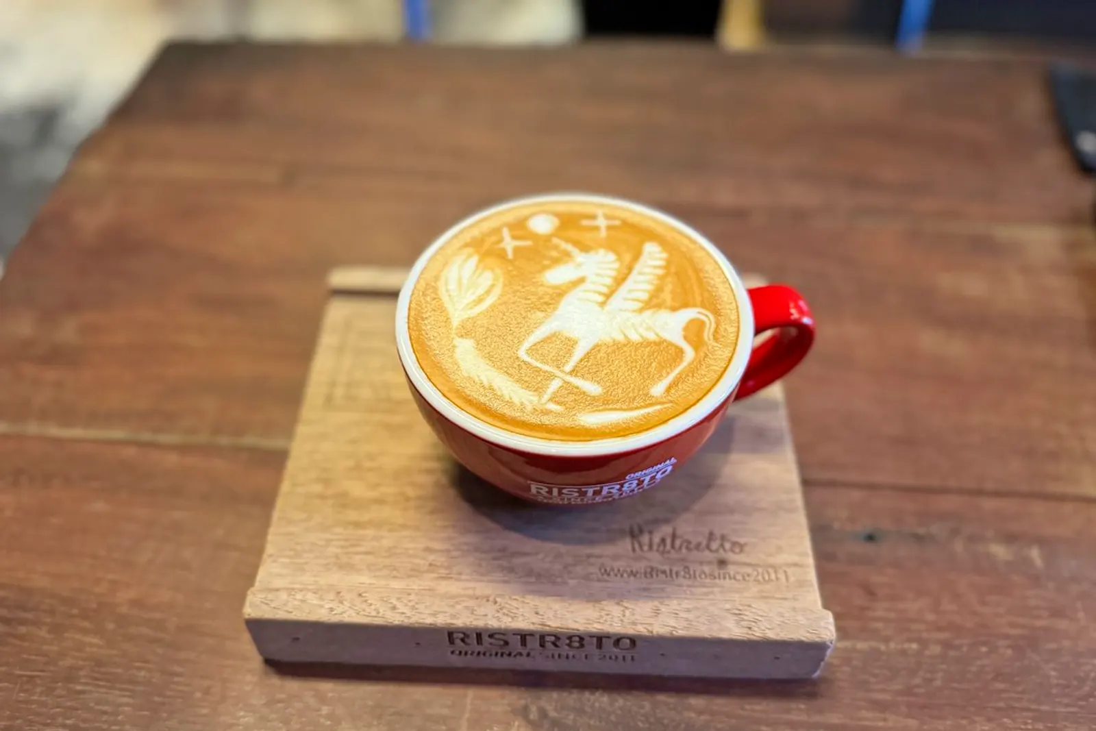 A latte with a winged horse poured in the foam, in a red Ristr8to cup on a wooden board