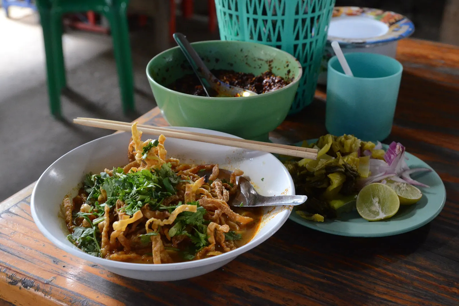 Khao soi with crispy noodles and coriander, with a bowl of roasted chilli paste and a plate of pickled mustard greens, red shallots and lime