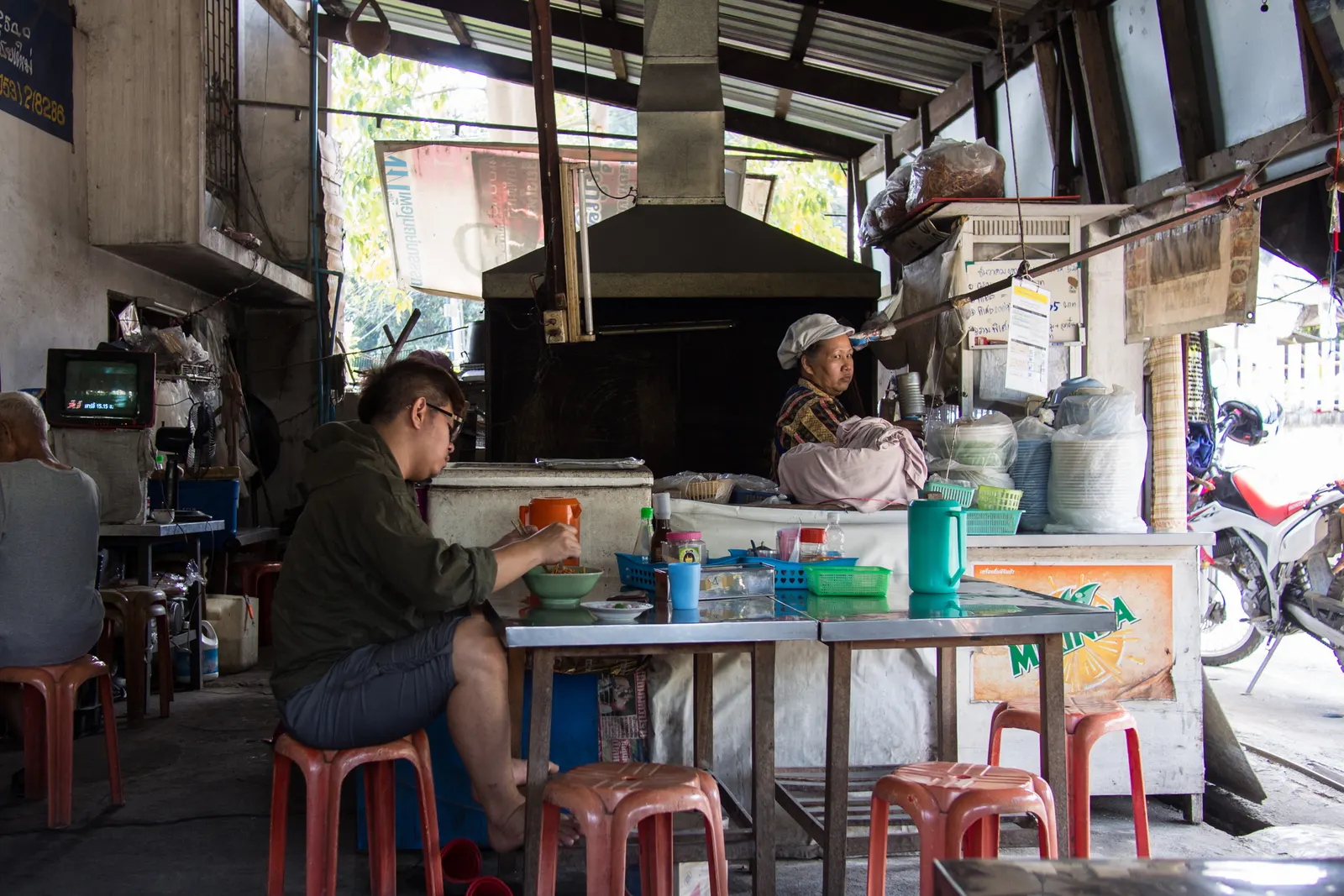 A diner at a steel table by the kitchen counter inside Khao Soi Mae Manee in Chiang Mai, photographed in 2014
