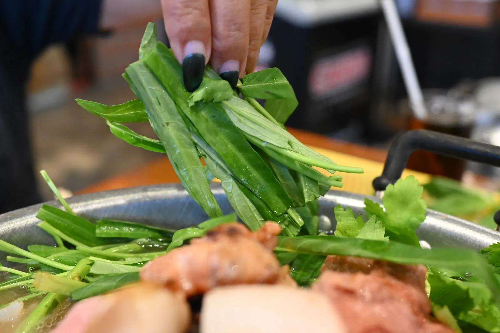 A hand dropping morning glory into the broth around a mookata grill, with pork cooking on the dome