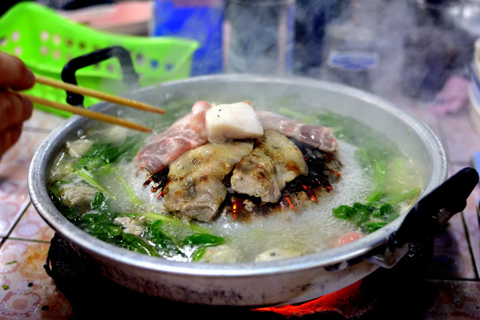 A mookata pan over glowing charcoal: a lump of pork fat on top of the dome, pork grilling around it and greens simmering in the broth moat