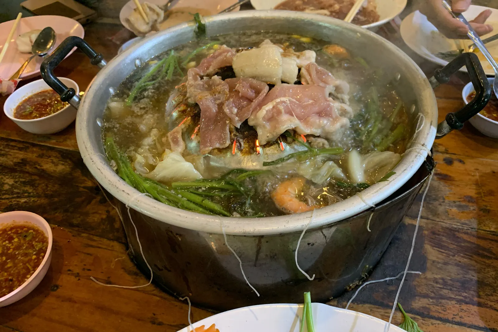 A mookata table: the pan full of pork, prawns and morning glory, with bowls of red dipping sauce and plates of raw meat around it