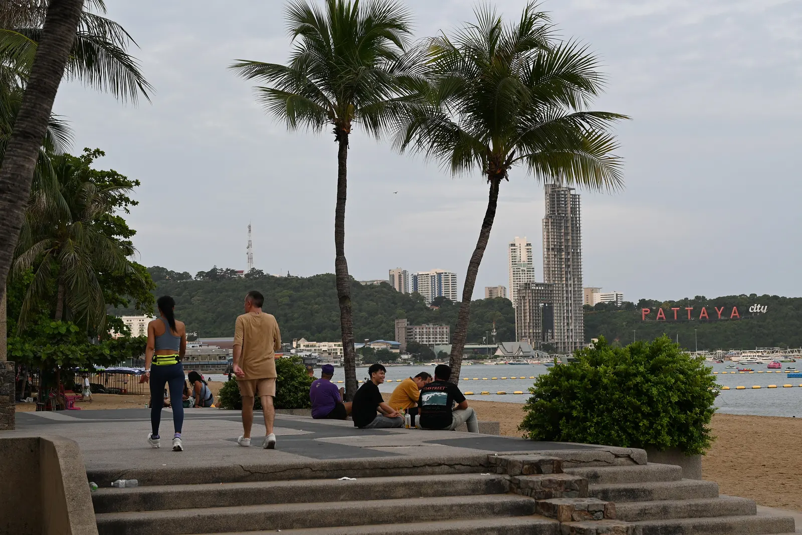 คนเดินใต้ต้นมะพร้าวบนทางเดินริมหาดพัทยา ไกล ๆ เป็นป้าย PATTAYA CITY สีแดงบนเขาพระตำหนัก