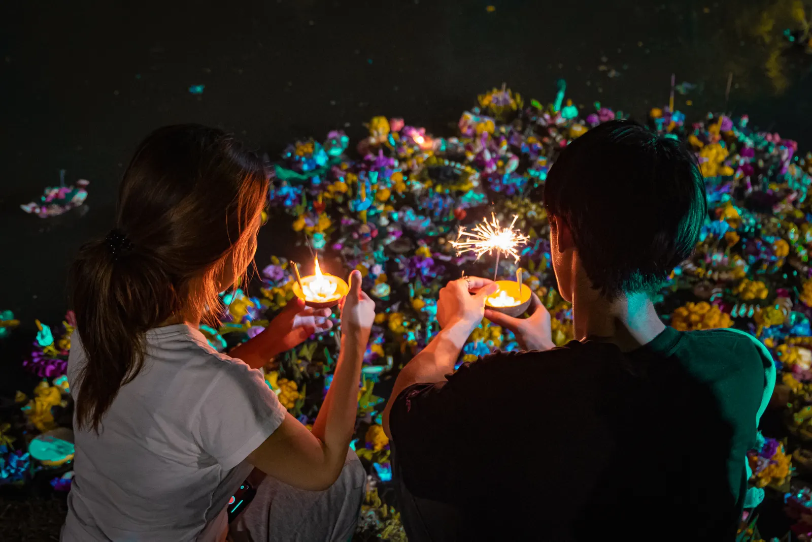 Two people lighting the candles on their krathongs at the water's edge on Loy Krathong night in November 2024